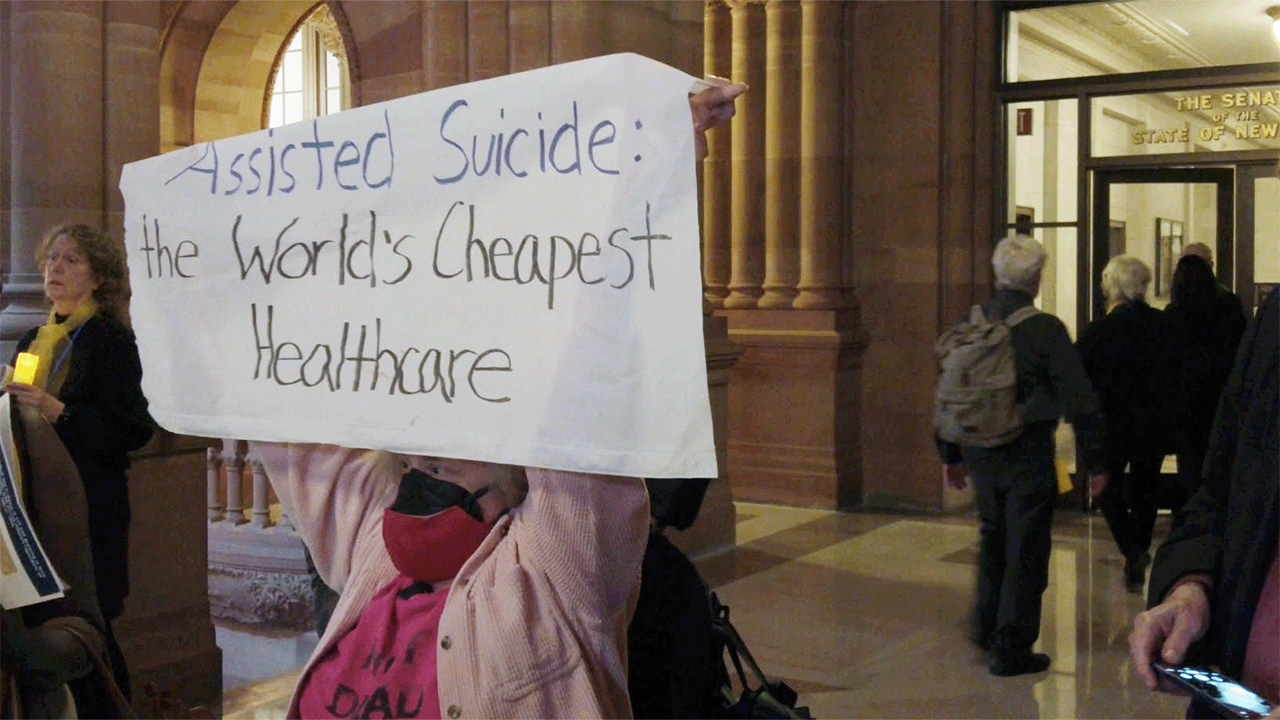 woman in government building holding sign that says "assisted suicide: The Worlds Cheapest Healthcare"